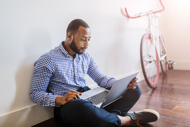Mann mit Laptop auf Holzboden sitzend mit Fahrrad im Hintergrund - GIOF03142