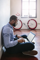 Man using laptop sitting on wooden floor with bicycle in background - GIOF03143