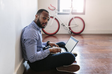 Man using laptop sitting on wooden floor with bicycle in background - GIOF03144