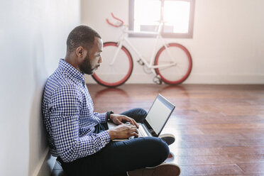 Man using laptop sitting on wooden floor with bicycle in background - GIOF03145