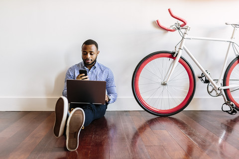 Man sitting on wooden floor with laptop and cell phone and bicycle next to him stock photo