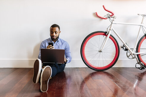 Mann sitzt auf Holzboden mit Laptop und Handy und Fahrrad neben ihm - GIOF03146