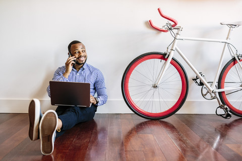 Smiling man on cell phone sitting on wooden floor with bicycle next to him stock photo