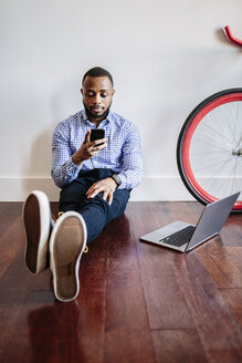 Mann sitzt auf Holzboden mit Laptop und Handy und Fahrrad neben ihm - GIOF03148