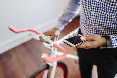 Nahaufnahme eines Mannes, der sein Telefon überprüft, während er ein Fahrrad im Haus hält - GIOF03150