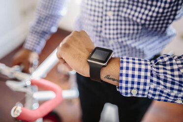 Nahaufnahme eines Mannes, der seine Smartwatch überprüft, während er ein Fahrrad in der Wohnung hält - GIOF03151