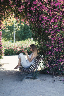 Frau mit Handy sitzt auf Bank im Park unter rosa Blüten - JPF00273