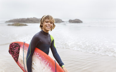 Spain, Aviles, portrait of smiling young surfer carrying surfboard on the beach - MGOF03563