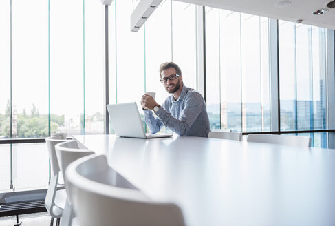 Mann mit Laptop und Tasse Kaffee sitzt am Konferenztisch im Büro - DIGF02724
