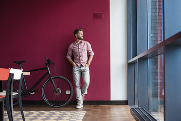 Man with bicycle standing in modern office looking out of window - DIGF02740