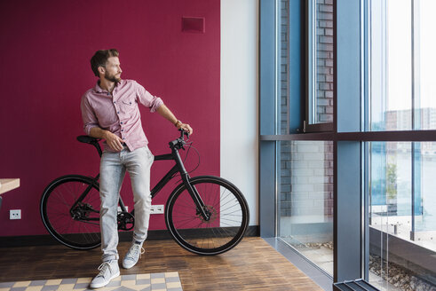 Mann mit Fahrrad steht in einem modernen Büro und schaut aus dem Fenster - DIGF02741