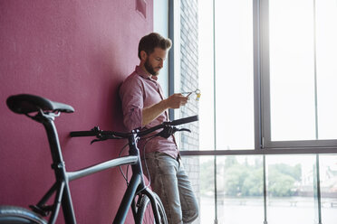 Mann mit Fahrrad steht in einem modernen Büro und schaut auf sein Handy - DIGF02742