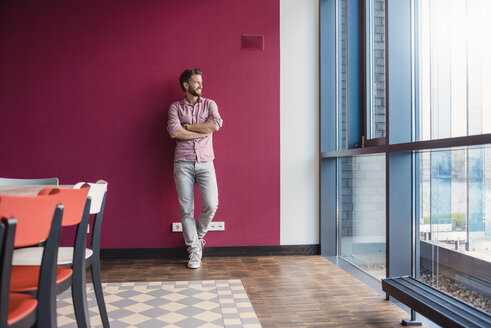 Mann lehnt an lila Wand in modernem Büro und schaut aus dem Fenster - DIGF02745
