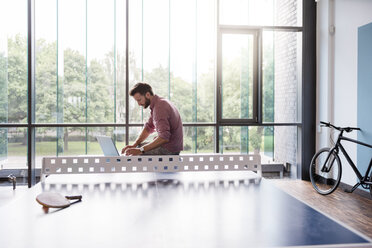 Mann benutzt Laptop im Pausenraum eines modernen Büros an einer Tischtennisplatte - DIGF02754