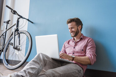 Laughing man using laptop sitting on the floor in office - DIGF02761