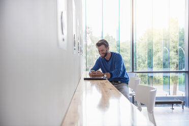 Lächelnder Mann schaut auf sein Handy auf dem Tisch am Fenster - DIGF02765