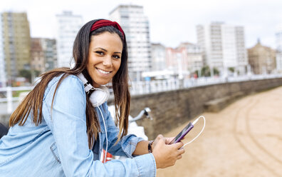 Spain, Gijon, smiling young woman with cell phone and headphones at waterfront promenade - MGOF03572