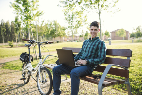 Young man with bicycle using laptop on park bench stock photo