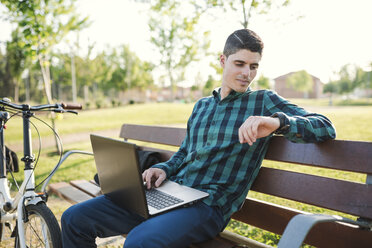 Junger Mann mit Laptop auf einer Parkbank, der die Zeit überprüft - RAEF01909