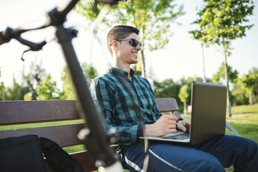 Young man with bicycle using laptop on park bench - RAEF01910