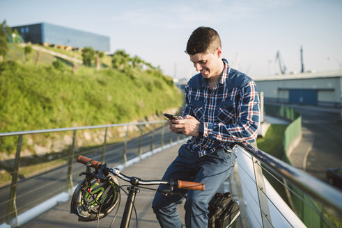 Lächelnder junger Mann mit Fahrrad, der auf sein Smartphone schaut - RAEF01915