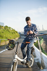 Lächelnder junger Mann mit Fahrrad, der auf sein Smartphone schaut - RAEF01916
