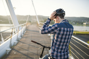 Young man on bicycle putting on helmet on a bridge - RAEF01917