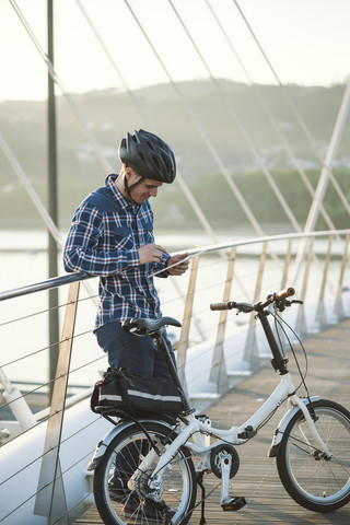 Junger Mann mit Klappfahrrad auf einer Brücke mit Blick auf ein Tablet, lizenzfreies Stockfoto