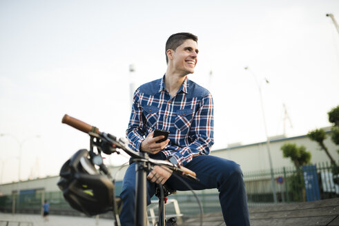 Glücklicher junger Mann mit Fahrrad und Mobiltelefon - RAEF01927