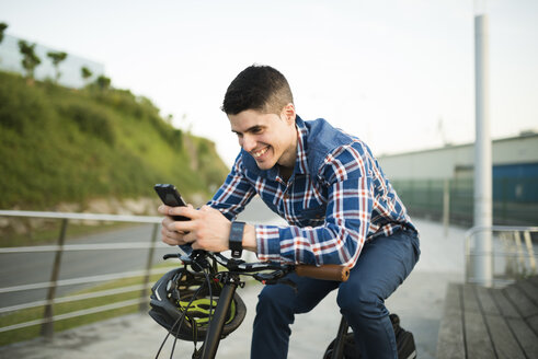 Lächelnder junger Mann mit Fahrrad, der auf sein Smartphone schaut - RAEF01929