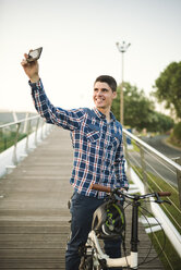 Lächelnder junger Mann mit Klapprad auf einer Brücke, der ein Selfie macht - RAEF01930