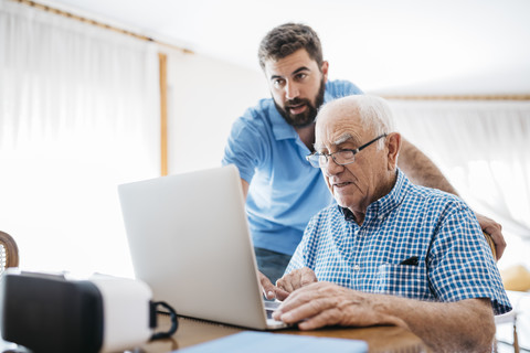 Erwachsener Enkel bringt seinem Großvater den Umgang mit dem Laptop bei, lizenzfreies Stockfoto