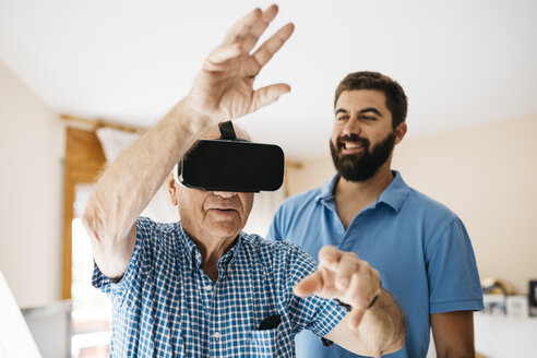 Ein älterer Mann benutzt eine Virtual-Reality-Brille zu Hause, während sein erwachsener Enkel ihn beobachtet - JRFF01424