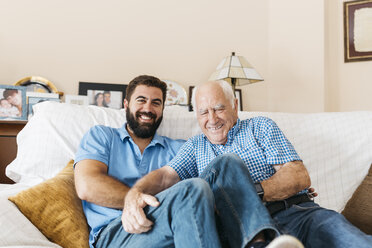Porträt eines erwachsenen Enkels und seines Großvaters, die zu Hause auf der Couch sitzen und sich gegenseitig kitzeln - JRFF01432