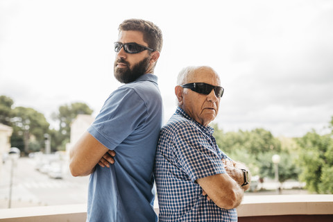 Portrait of adult grandson and his grandfather wearing sunglasses standing back to back on balcony stock photo