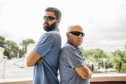 Portrait of adult grandson and his grandfather wearing sunglasses standing back to back on balcony - JRFF01438