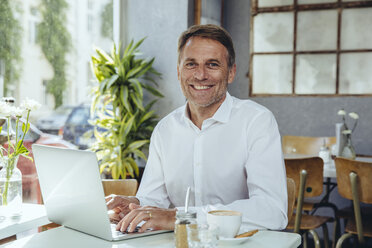 Portrait of smiling businessman working in cafe with laptop - MFF03856