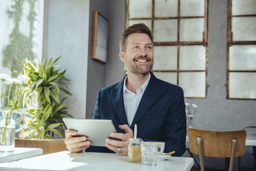 Smiling businessman with tablet in a cafe looking up - MFF03860