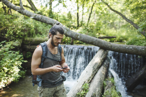 Junger Mann hält Mobiltelefon an einem Wasserfall im Wald - VPIF00028