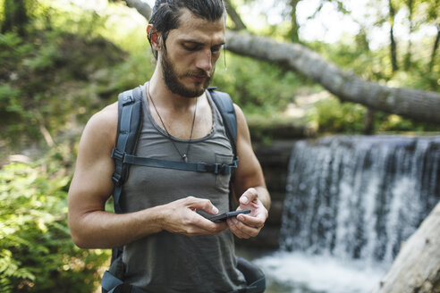 Junger Mann hält Mobiltelefon an einem Wasserfall im Wald - VPIF00029