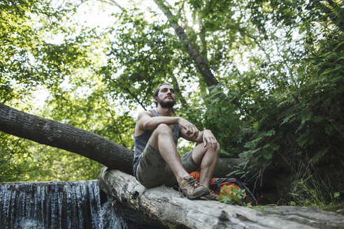 Junger Mann sitzt auf einem Baumstamm an einem Wasserfall im Wald - VPIF00031