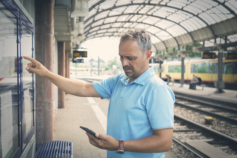 Mann mit Smartphone steht auf dem Bahnsteig vor einem Fahrplan, lizenzfreies Stockfoto