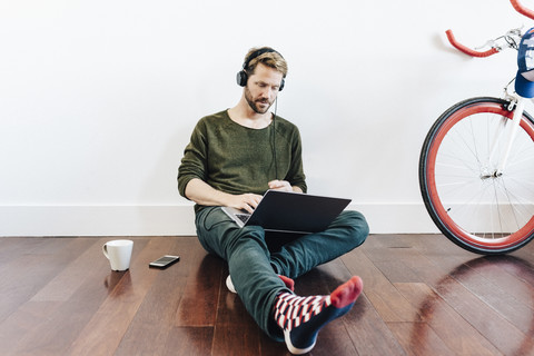 Mann mit Kopfhörern, der zu Hause auf dem Boden sitzt und einen Laptop benutzt, lizenzfreies Stockfoto