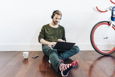 Mann mit Kopfhörern, der zu Hause auf dem Boden sitzt und einen Laptop benutzt - GIOF03161