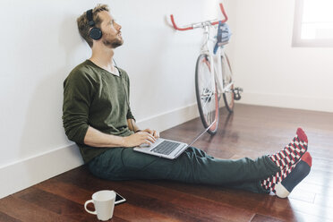 Nachdenklicher Mann mit Kopfhörern und Laptop, der zu Hause auf dem Boden sitzt - GIOF03163
