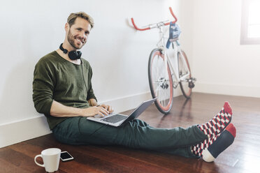 Porträt eines lächelnden Mannes mit Kopfhörern, der zu Hause auf dem Boden sitzt und einen Laptop benutzt - GIOF03166