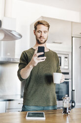 Porträt eines Mannes, der mit einer Tasse Kaffee in der Küche steht und ein Selfie mit seinem Smartphone macht - GIOF03171