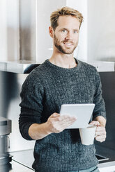 Portrait of smiling man standing in kitchen with tablet and coffee mug - GIOF03174