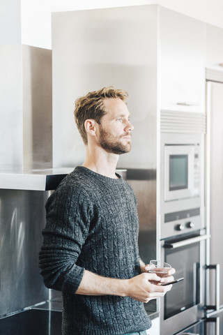 Nachdenklicher Mann in der Küche mit Mobiltelefon und einem Glas Wasser, lizenzfreies Stockfoto