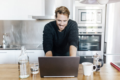 Porträt eines lächelnden Mannes, der in der Küche steht und einen Laptop benutzt - GIOF03177